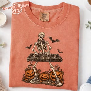 May include: A coral-coloured t-shirt featuring a graphic of a skeleton wearing headphones and DJing on a turntable console. The design includes three jack-o'-lantern pumpkins at the base and three flying bats. The shirt has a Comfort Colors label.