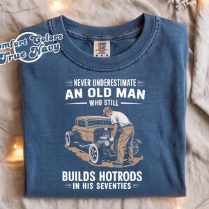 May include: True Navy Comfort Colors t-shirt with the text "NEVER UNDERESTIMATE AN OLD MAN WHO STILL BUILDS HOTRODS IN HIS SEVENTIES." The graphic shows a vintage car and a man working on it.