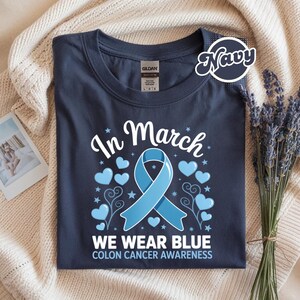 May include: Navy blue t-shirt with the text "In March We Wear Blue Colon Cancer Awareness". The design includes a light blue ribbon, hearts, and stars. The shirt is folded on a cream-colored blanket.