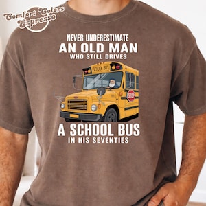 Op de afbeelding: Bruin t-shirt met de tekst "NEVER UNDERESTIMATE AN OLD MAN WHO STILL DRIVES A SCHOOL BUS IN HIS SEVENTIES." Een gele schoolbus staat op de voorkant.