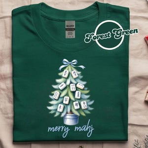 Könnte beinhalten: Forstgrünes T-Shirt mit einem Weihnachtsbaum-Design aus Mahjong-Steinen. Der Baum ist mit einer blauen Schleife verziert und steht in einem silbernen Topf. Die Worte "merry mahj" sind in einer Schreibschrift geschrieben.