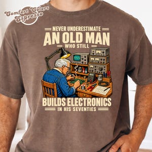 Op de afbeelding: Een bruin t-shirt met een afbeelding van een persoon die aan een bureau met elektronische apparatuur werkt. De tekst luidt: "NEVER UNDERESTIMATE AN OLD MAN WHO STILL BUILDS ELECTRONICS IN HIS SEVENTIES" en "Comfort Colors Espresso".