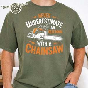 Op de afbeelding: Olijfgroen t-shirt met de tekst "Never Underestimate an Old Man with a Chainsaw" in wit en oranje. De afbeelding bevat een kettingzaag illustratie. Het shirt heeft een ronde hals en korte mouwen.