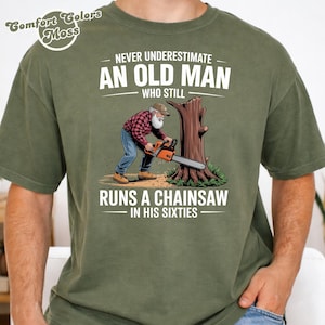 May include: An olive green t-shirt featuring the text "NEVER UNDERESTIMATE AN OLD MAN WHO STILL RUNS A CHAINSAW IN HIS SIXTIES." The graphic depicts an elderly man using a chainsaw. The shirt also has the Comfort Colors logo.