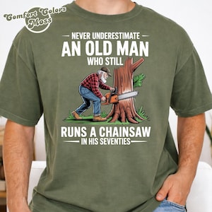 May include: Olive green t-shirt with a graphic of an older man using a chainsaw on a tree. Text reads "NEVER UNDERESTIMATE AN OLD MAN WHO STILL RUNS A CHAINSAW IN HIS SEVENTIES." The shirt has a Comfort Colors Moss label.