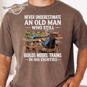 May include: A brown t-shirt featuring a graphic of an older person working on a model train set at a workbench. The text reads: NEVER UNDERESTIMATE AN OLD MAN WHO STILL BUILDS MODEL TRAINS IN HIS EIGHTIES. The Comfort Colors Espresso logo is visible.