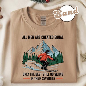 Op de afbeelding: Zandkleurig T-shirt met de tekst "ALL MEN ARE CREATED EQUAL" boven een afbeelding van een skiër en de tekst "ONLY THE BEST STILL GO SKIING IN THEIR SEVENTIES". De skiër draagt een rode jas en een zwarte broek.
