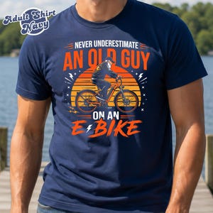 Puede incluir: Una camiseta azul marino con un gráfico de una persona con barba montando una bicicleta eléctrica frente a un sol naranja. El texto dice: NEVER UNDERESTIMATE AN OLD GUY ON AN E-BIKE. La esquina superior izquierda incluye: Adult Shirt Navy.