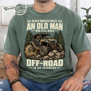 Op de afbeelding: Een mosgroen t-shirt met een afbeelding van een terreinwagen die over rotsen en bomen rijdt. De tekst luidt: NEVER UNDERESTIMATE AN OLD MAN WHO STILL RIDES OFF-ROAD IN HIS SEVENTIES. Het t-shirt heeft een ronde hals en korte mouwen.