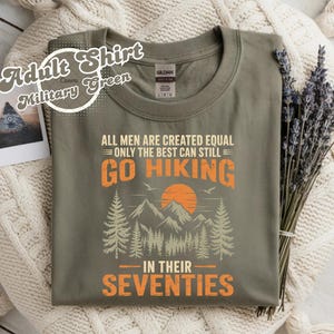 Puede incluir: Camiseta para adulto verde militar con el texto "ALL MEN ARE CREATED EQUAL ONLY THE BEST CAN STILL GO HIKING IN THEIR SEVENTIES" en naranja y blanco. El diseño incluye una escena de montaña con árboles y un sol naciente.