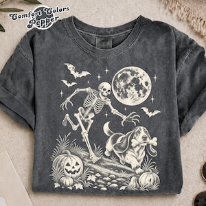 以下が含まれることがあります： チャコールグレーのTシャツで、骨を口にくわえたバセットハウンドの横を走る骸骨のクリーム色のグラフィックが描かれています。満月、コウモリ、草地に置かれた2つのジャック・オー・ランタンがデザインされています。