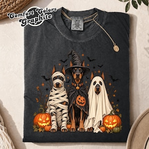 May include: A charcoal gray t-shirt featuring a graphic of three Doberman dogs dressed for Halloween. One dog is wrapped in bandages like a mummy, one wears a witch hat and pumpkin cape, and one is covered by a white ghost sheet, with pumpkins.
