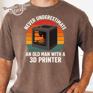 Op de afbeelding: Een bruin t-shirt met een afbeelding van een 3D-printer in een zonsondergangcirkel. De tekst luidt "NEVER UNDERESTIMATE AN OLD MAN WITH A 3D PRINTER" in witte hoofdletters. In de linkerbovenhoek staat de tekst "Comfort Colors Espresso".