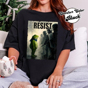 May include: Black comfort color t-shirt featuring a graphic of a green frog wearing a blue scarf, standing before a line of riot police. The word "RESIST" is printed above the frog. The shirt is black.