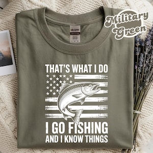 May include: Military green t-shirt with a graphic of a fish and an American flag design. The text reads, "THAT'S WHAT I DO, I GO FISHING AND I KNOW THINGS." A small photo and dried flowers are next to the shirt.