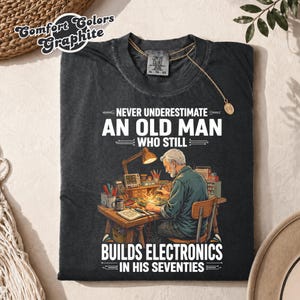 May include: A charcoal gray t-shirt featuring a graphic of a person working at a desk with electronic tools and a lamp. The text reads: NEVER UNDERESTIMATE AN OLD MAN WHO STILL BUILDS ELECTRONICS IN HIS SEVENTIES. The shirt has a Comfort Colors label.