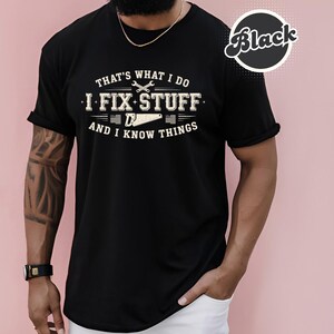 May include: Black t-shirt with the text "THAT'S WHAT I DO I FIX STUFF AND I KNOW THINGS" in a distressed, vintage style. The design includes tools like a wrench and saw. The shirt has rolled-up sleeves.