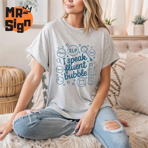 May include: A gray t-shirt with a blue graphic design that says "I speak fluent bubble" with a speech bubble and other speech bubbles that say "More", "Again", "SLP", "Yep", and "Your Turn".