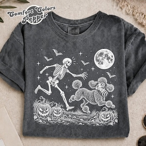 May include: A charcoal grey crew neck t-shirt featuring a white graphic print of a skeleton running alongside a poodle. The design includes a full moon, stars, bats, and two jack-o'-lanterns. The text Comfort Colors Pepper is printed in the corner.