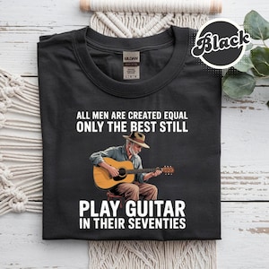 Könnte beinhalten: Schwarzes T-Shirt mit dem Text "ALL MEN ARE CREATED EQUAL ONLY THE BEST STILL PLAY GUITAR IN THEIR SEVENTIES." Das Shirt zeigt eine Illustration eines Mannes, der Gitarre spielt. Das Gildan-Markenetikett ist sichtbar.