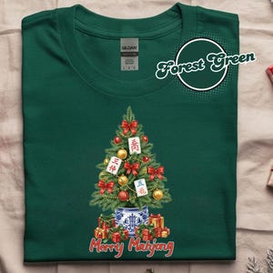 May include: Forest green t-shirt featuring a Christmas tree design with Mahjong tiles, red bows, and gold ornaments. The tree is in a blue and white vase, with wrapped gifts at the base. The text "Merry Mahjong" is at the bottom.