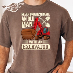 May include: A brown short-sleeve t-shirt featuring a graphic of a red excavator and two crossed shovels. The text reads: NEVER UNDERESTIMATE AN OLD MAN WITH AN EXCAVATOR. The top left corner includes the text: Comfort Colors Espresso.