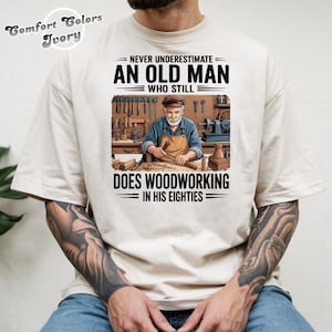 Op de afbeelding: Ivoor Comfort Colors T-shirt met de tekst "NEVER UNDERESTIMATE AN OLD MAN WHO STILL DOES WOODWORKING IN HIS EIGHTIES." De afbeelding toont een houtbewerker in zijn werkplaats. Het shirt heeft een relaxte pasvorm.
