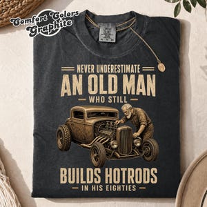 Pode incluir: Uma t-shirt cinza grafite com um gráfico de uma pessoa idosa a trabalhar num carro hot rod vintage. O texto diz: NEVER UNDERESTIMATE AN OLD MAN WHO STILL BUILDS HOTRODS IN HIS EIGHTIES. A t-shirt tem uma etiqueta de pescoço Comfort Colors.