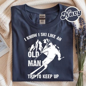 Könnte beinhalten: Marineblaues T-Shirt mit weißer Grafik eines Skifahrers und dem Text "I KNOW I SKI LIKE AN OLD MAN TRY TO KEEP UP". Das Shirt liegt gefaltet auf einer cremefarbenen Decke.