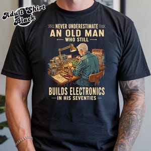 May include: A black t-shirt featuring a graphic illustration of a person working at a desk with electronic components and tools. The text reads: NEVER UNDERESTIMATE AN OLD MAN WHO STILL BUILDS ELECTRONICS IN HIS SEVENTIES. The design includes a lamp.