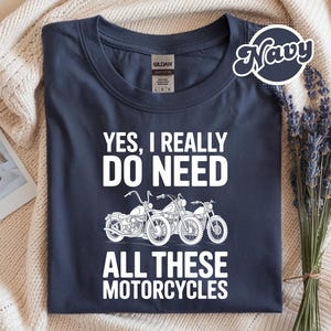 Può includere: T-shirt blu navy con la scritta bianca "YES, I REALLY DO NEED ALL THESE MOTORCYCLES". La grafica presenta tre illustrazioni di motociclette. La maglietta è piegata su una superficie color crema.