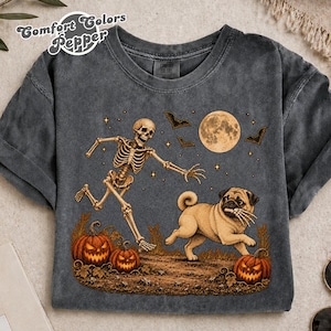 Puede incluir: Una camiseta gris carbón con un gráfico de un esqueleto corriendo tras un pug beige que sostiene una mano esquelética en su boca. La escena incluye dos calabazas de Halloween, una luna llena, murciélagos y estrellas. El texto Comfort Colors Pepper es visible.
