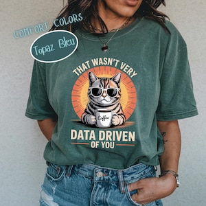 May include: Green t-shirt featuring a cat in sunglasses holding a coffee mug, with the text "THAT WASN'T VERY DATA DRIVEN OF YOU." The shirt also displays "COMFORT COLORS" and "Topaz Bleu". The t-shirt is a comfortable fit.