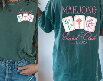 Puede incluir: Camiseta verde azulado con un diseño de Mahjong Social Club. La parte delantera muestra el nombre "Isabella" con tres fichas de Mahjong. La parte trasera muestra "Mahjong Social Club", el año "EST 2025" y tres fichas de Mahjong.