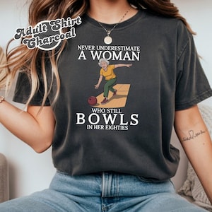 Op de afbeelding: Antracietgrijs t-shirt met de tekst "NEVER UNDERESTIMATE A WOMAN WHO STILL BOWLS IN HER EIGHTIES." De afbeelding toont een cartoon van een vrouw die aan het bowlen is. De tekst "Adult Shirt Charcoal" staat in een cirkel bovenaan.
