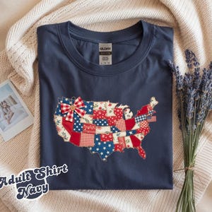 Puede incluir: Camiseta azul marino para adultos con un diseño de patchwork del mapa de Estados Unidos. El mapa presenta patrones rojos, blancos y azules. Un pequeño lazo está en la parte superior. El texto "Adult Shirt Navy" está en la parte inferior.
