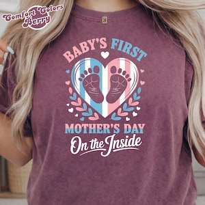 Può includere: T-shirt bordeaux con la scritta "Baby's First Mother's Day On the Inside". Il design include un cuore con impronte di bambino, strisce blu e rosa e accenti floreali. La maglietta ha uno scollo tondo.