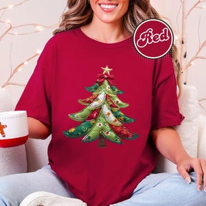 Op de afbeelding: Rood T-shirt met een kerstboomontwerp. De boom is versierd met groene, rode en witte stofpatronen, bekroond met een gouden ster en een rode strik. Een witte mok met een peperkoekmannetje wordt vastgehouden.