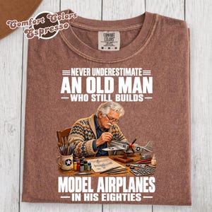 Peut inclure: Un t-shirt marron avec une illustration représentant une personne âgée aux cheveux blancs et à lunettes, portant un gilet à motifs tout en construisant un modèle réduit d'avion. Le texte indique : NEVER UNDERESTIMATE AN OLD MAN WHO STILL BUILDS MODEL AIRPLANES IN HIS EIGHTIES.