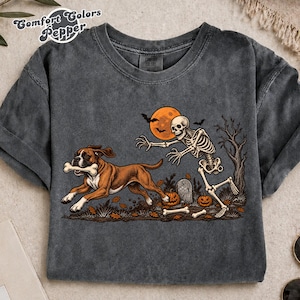 May include: A charcoal gray crew neck t-shirt featuring a graphic of a brown boxer dog running with a bone in its mouth, pursued by a skeleton. The scene includes a full orange moon, bats, pumpkins, and a tombstone in a dark, autumnal setting.