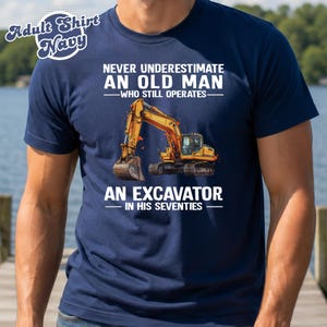 Może przedstawiać: Granatowy t-shirt z krótkim rękawem z grafiką żółtej koparki. Napis głosi: NEVER UNDERESTIMATE AN OLD MAN WHO STILL OPERATES AN EXCAVATOR IN HIS SEVENTIES. Koszulka posiada okrągły dekolt oraz standardowy krój.
