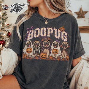Puede incluir: Una camiseta gris carbón con el texto BOOPUG en blanco con lunares naranjas. El gráfico muestra cinco carlinos con disfraces de Halloween, incluyendo fantasmas, un esqueleto, una bruja y un vampiro, rodeados de calabazas y hojas de otoño.