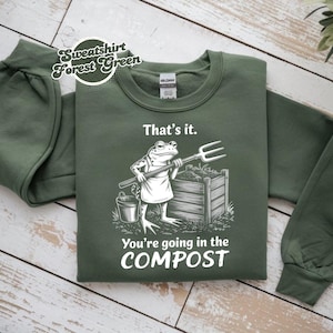 Op de afbeelding: Bosgroene sweatshirt met de tekst "That's it. You're going in the COMPOST" en een kikker met een hooivork. De sweatshirt heeft lange mouwen en een ronde hals.