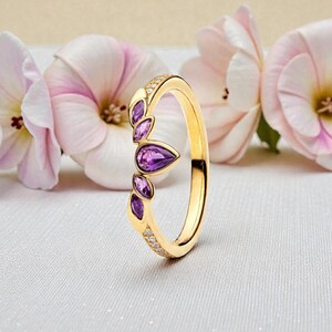 May include: A gold ring featuring a leaf-shaped design, embellished with purple gemstones and tiny clear stones. The ring is set against a backdrop of pink and white flowers, creating a delicate and elegant aesthetic.