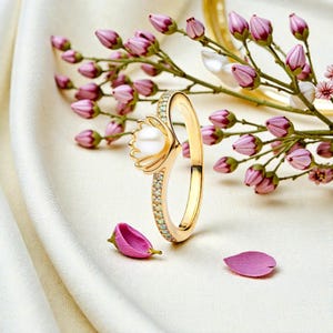 Könnte beinhalten: Ein goldener Ring mit einer Perle in der Mitte und kleinen, funkelnden Steinen entlang des Bandes. Der Ring wird vor dem Hintergrund von rosa Blütenknospen und Blütenblättern präsentiert, was eine zarte und elegante Ästhetik erzeugt.