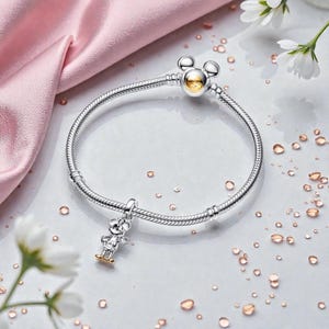 May include: A silver bracelet with a Mickey Mouse charm and clasp. The charm features a small Mickey Mouse figure with gold-colored accents. The clasp has a Mickey Mouse head design with a gold-colored center. The bracelet is on a white surface with flower accents.