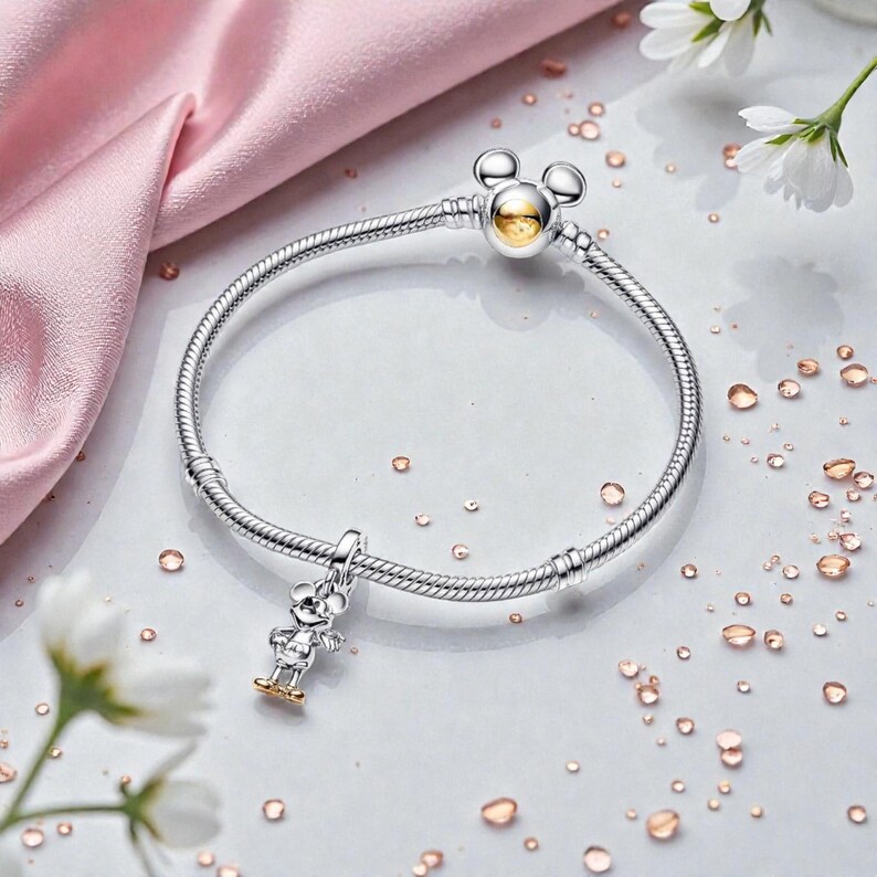 May include: A silver bracelet with a Mickey Mouse charm and clasp. The charm features a small Mickey Mouse figure with gold-colored accents. The clasp has a Mickey Mouse head design with a gold-colored center. The bracelet is on a white surface with flower accents.