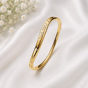 May include: A gold hinged bangle bracelet featuring a curved, asymmetrical design. The surface is polished and accented with a cluster of small, clear, round-cut stones embedded along one side, creating a sparkling, constellation-like pattern.
