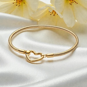 May include: Gold-colored bracelet with a heart-shaped clasp. The bracelet has a snake chain design and is displayed on a white satin surface. The heart clasp adds a romantic touch to the jewelry.