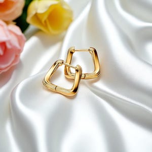 May include: A pair of gold-colored, square hoop earrings. The earrings are displayed on a white, silky fabric. The earrings have a geometric design and a polished finish. The background includes pink and yellow flowers.